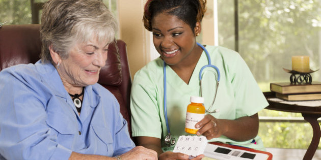 Caring, African descent home healthcare nurse conducts medical consultation with senior adult woman at her home or nursing home.  The female patient is sitting in her comfortable chair while reviewing her medications information with her nurse or doctor.  Backyard can be seen through the windows.